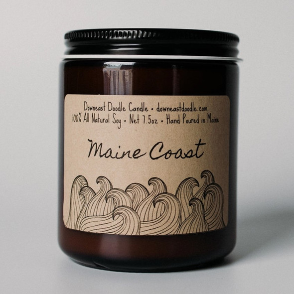 a candle in an amber glass jar with black lid and kraft label with handwriting style graphics that read "Maine Coast" above playful doodle style line drawings of ocean waves