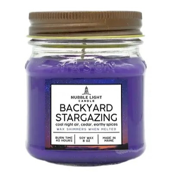 a candle in a clear glass mason jar with purple wax and a label that reads "Backyard Stargazing" against a white background